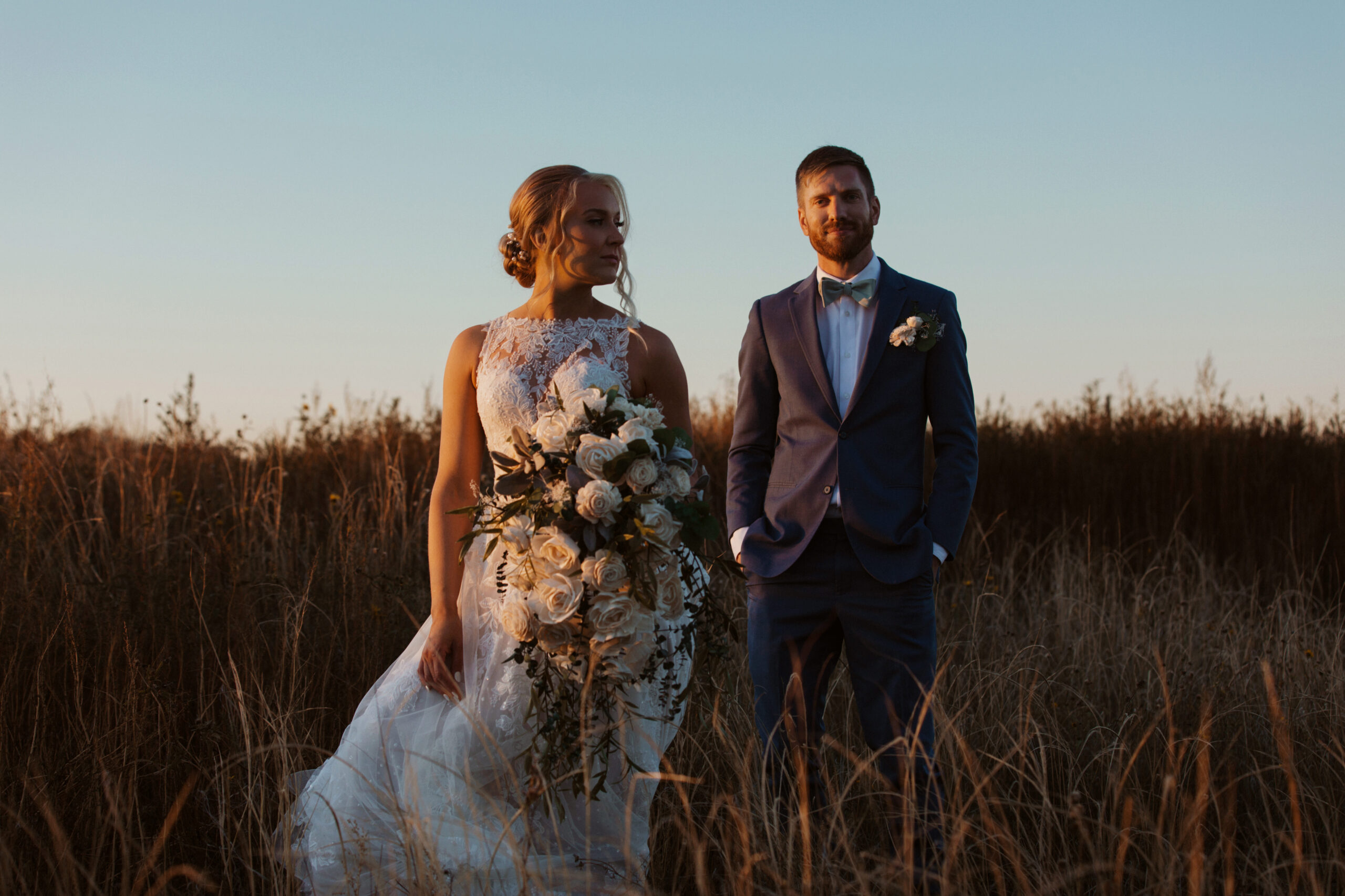 Bride and groom stand in a field at dusk in an editorial pose.